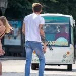 Фотография мужчины с татуировками на руках и спине, стоящего на улице. Современный стиль, городская обстановка, повседневная одежда. Детализированное фото тату.