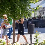 Фотография мужчины с татуировкой на руке, идущего по улице в окружении людей и зелени.