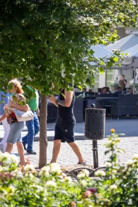Фотография мужчины с татуировкой на руке, идущего по улице в окружении людей и зелени.