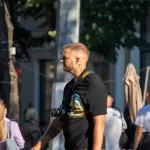Мужчина с бородой, одетый в черную футболку, стоит на улице с фотоаппаратом. На руке видна татуировка в стиле графики.