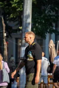 Мужчина с бородой, одетый в черную футболку, стоит на улице с фотоаппаратом. На руке видна татуировка в стиле графики.