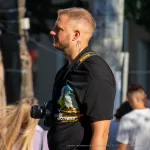Мужчина с бородой, одетый в черную футболку, смотрит в видоискатель фотоаппарата. На руке видна татуировка в стиле графики.