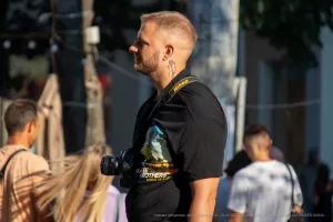 Мужчина с бородой, одетый в черную футболку, смотрит в видоискатель фотоаппарата. На руке видна татуировка в стиле графики.