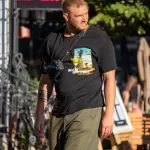 Мужчина с бородой, одетый в черную футболку и шорты, идет по улице с фотоаппаратом. На руке видна татуировка в стиле графики.