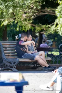 Фотография пары, сидящей на скамейке в парке, с акцентом на татуировку на руке парня. Солнечный день и спокойная атмосфера.