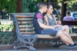 Фотография пары, сидящей на скамейке в парке, с акцентом на татуировку на руке парня. Солнечный день и спокойная атмосфера.