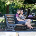 Фотография пары, сидящей на скамейке в парке, с акцентом на татуировку на руке парня. Солнечный день и спокойная атмосфера.