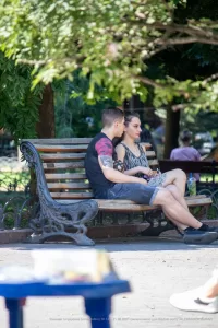 Фотография пары, сидящей на скамейке в парке, с акцентом на татуировку на руке парня. Солнечный день и спокойная атмосфера.