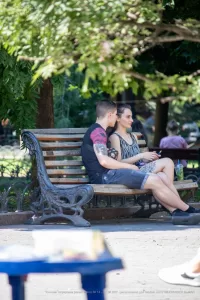 Фотография пары, сидящей на скамейке в парке, с акцентом на татуировку на руке парня. Солнечный день и спокойная атмосфера.