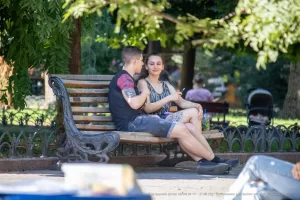 Фотография пары, сидящей на скамейке в парке, с акцентом на татуировку на руке парня. Солнечный день и спокойная атмосфера.