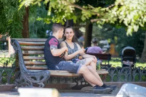 Фотография пары, сидящей на скамейке в парке, с акцентом на татуировку на руке парня. Яркий солнечный день и расслабленная атмосфера.