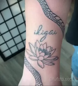 Фото татуировки лотоса с надписью 'ikigai' и изображением змеи на руке. Черно-белый рисунок в графическом стиле.