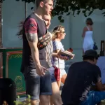 Фотография мужчины с татуировкой на руке, идущего по улице. На заднем плане видны другие люди и городская среда.