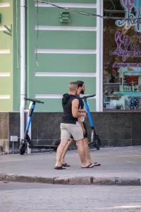 Фото мужчины с татуировкой на руке, стоящего рядом с электросамокатом. Рисунок татуировки выполнен в черных тонах.