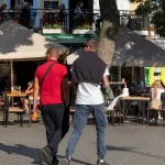 Фотография мужчины с татуировкой на руке, идущего по улице города. Татуировка выполнена в темных тонах и видна на предплечье.