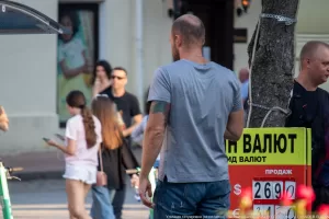 Фото тату на руке мужчины, изображающее темный рисунок. Татуировка выполнена в стиле графика и покрывает часть предплечья. Мужчина стоит возле обменного пункта.