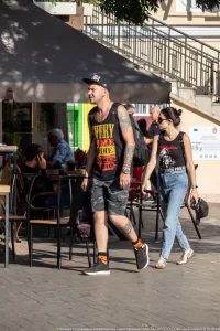 Фотография мужчины с татуировками на руках и ногах, одетого в майку и шорты, идущего по улице в кепке.
