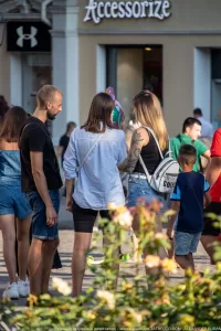 Фото тату на плече женщины, изображающее темные узоры и растительные мотивы. Женщина стоит на улице, разговаривая с другими людьми.