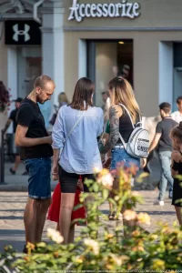 Фото тату на плече женщины, изображающее темные узоры и растительные мотивы. Женщина стоит на улице, разговаривая с другими людьми.