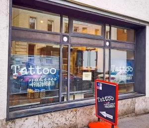Фасад тату-студии в Германии с большими витринами, надписью 'Tattoo' и рекламным щитом с контактной информацией.
