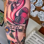Фото тату на руке: розовый фламинго с надписью "CREW" на ленте, выполненный в стиле олдскул.
