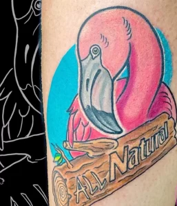 Фото татуировки головы фламинго на фоне голубого круга с надписью 'All Natural'. Яркие розовые и голубые цвета, детализированный рисунок.
