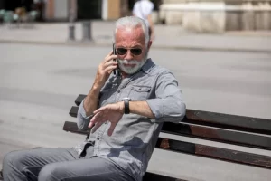 Фото пожилого мужчины в солнечных очках, разговаривающего по телефону на скамейке в городе, демонстрирующего активный образ жизни.