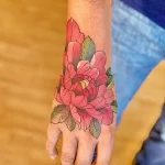 Фото тату с изображением пиона на тыльной стороне кисти руки. Цветная татуировка с использованием красных, зеленых и коричневых оттенков.