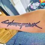Фото тату на руке: минималистичная надпись 'Cyberpunk', выполненная в черном цвете с использованием тонких линий и точечной техники.