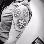 Фото тату: черно-белая татуировка с изображением снежинок и фразы "Winter is coming" из "Игры престолов" на плече. Минималистичный рисунок татуировки.