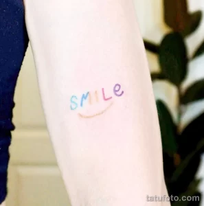 Фото тату: разноцветная надпись "SMILE" и улыбка, нанесенные на руку. Яркий и позитивный рисунок, поднимающий настроение.