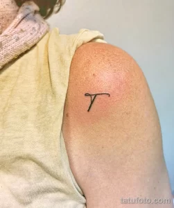 Фото тату: Маленькая черная татуировка буквы 'T', расположенная на плече. Минималистичный и лаконичный дизайн.