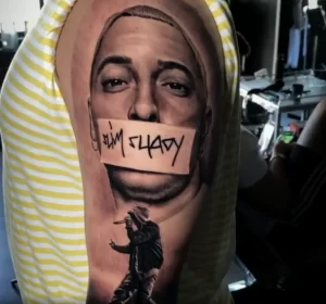 Фото татуировки с реалистичным портретом Эминема на плече, с надписью 'Slim Shady' на губах. Высокая детализация и реалистичная цветопередача.