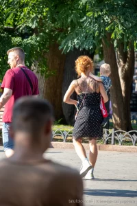 Фото тату на спине женщины с рыжими волосами, несущей ребенка. Рисунок татуировки представляет собой сложный цветочный узор с темными оттенками.