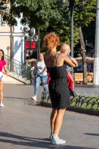Фото тату на спине рыжеволосой женщины, идущей по улице с ребенком. Рисунок татуировки выполнен в темных тонах, с преобладанием цветочных мотивов.