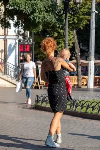 Фото тату на спине рыжеволосой женщины, держащей ребенка. Рисунок татуировки выполнен в темных тонах, с акцентом на цветочные мотивы.