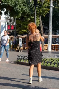 Фото тату на спине женщины, идущей по улице с ребенком. Рисунок татуировки выполнен в темных тонах, с элементами цветов и узоров.