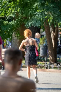 Фото тату на спине женщины с рыжими волосами, несущей ребенка. Рисунок татуировки представляет собой сложный цветочный узор с темными оттенками.