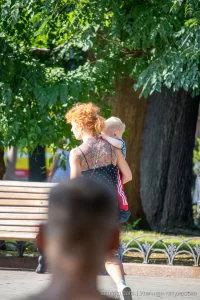 Фото тату на спине женщины с рыжими волосами, несущей ребенка. Рисунок татуировки представляет собой сложный цветочный узор с темными оттенками.