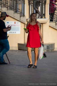 Фото тату на руке женщины в красном платье, идущей по улице. Рисунок татуировки выполнен в темных тонах и частично скрыт одеждой.