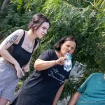Фото тату на плече девушки, сидящей на улице. Черно-белый рисунок татуировки в виде растительного орнамента, выполненный в реалистичном стиле.