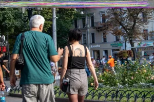 Фото тату на плече девушки, идущей по улице. Черно-белый рисунок татуировки в виде растительного орнамента, выполненный в реалистичном стиле.