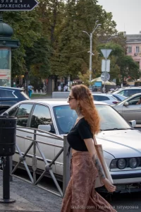 Фотография женщины с длинными рыжими волосами, идущей по улице с татуировкой на левой руке. Татуировка выполнена в темных тонах.
