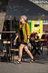 Фото тату на лодыжке женщины, изображающее темный рисунок, вероятно, растительный орнамент. Женщина идет по улице в желтой рубашке и шортах.