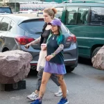 Фотография девушки с татуировками на руке и ноге, идущей по улице. Видны яркие цвета и детализированные рисунки в стиле олдскул и акварель.