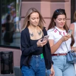Фото девушки с тату птиц на груди, смотрящей в телефон. Рисунок татуировки выполнен в черном цвете.