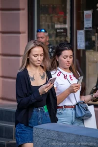 Фото двух девушек, одна из которых имеет тату птиц на груди, обе смотрят в телефоны. Рисунок татуировки выполнен в черном цвете.