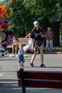 Фотография девушки с татуировкой на ноге на фоне воздушных шаров. На заднем плане городской пейзаж и рекламный баннер.