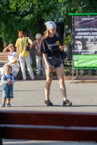 Фотография девушки, смотрящей в телефон, с видимой татуировкой на ноге. На заднем плане городской пейзаж и рекламный баннер.