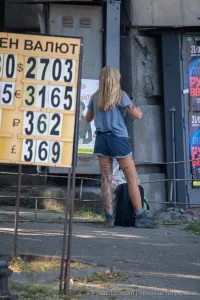 Фотография девушки с татуировкой на ноге, стоящей у входа в здание. Татуировка изображает цветы и надпись. Девушка одета в шорты и футболку.
